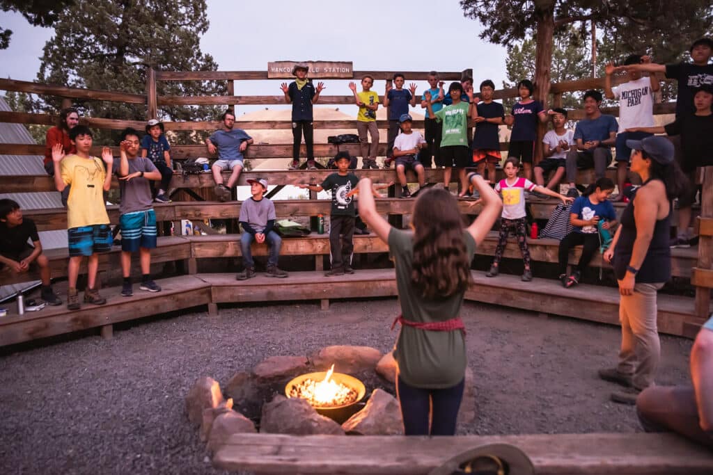 A group of kids sitting on wooden bleachers near a campfire.