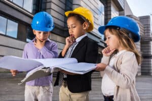Three children wearing construction hats, looking at building plans with curious expressions