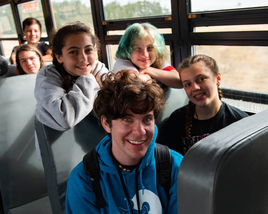Four kids sitting in rows on a school bus with their backpacks on and smiling