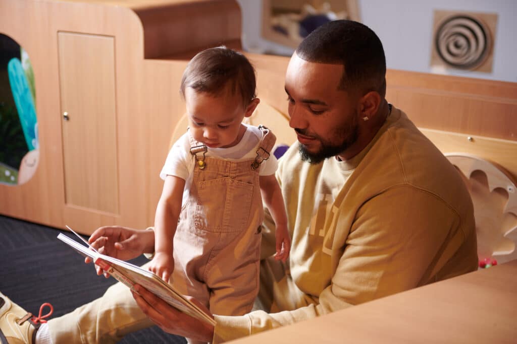 A man in a tan outfit reads a book with a toddler in overalls, sitting on the floor in OMSI's Science Playground.
