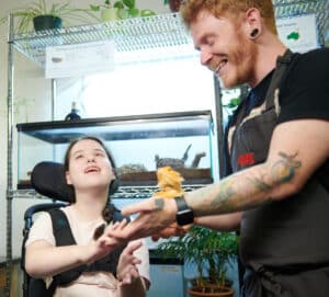 Staff member holding a lizard to a girl's hand sitting in a wheelchair