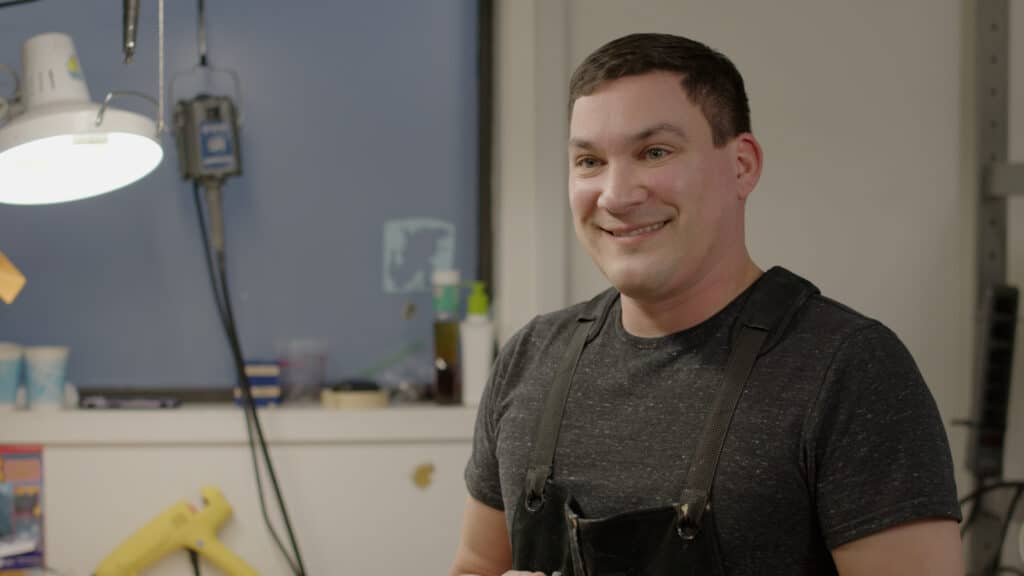 Photo of a man wearing a grey shirt and apron smiling during an interview