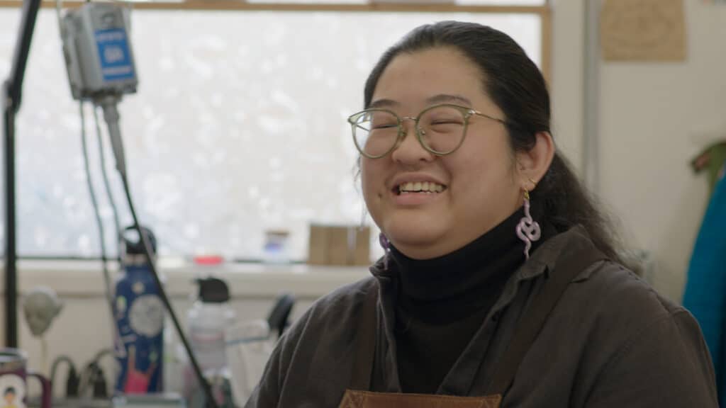 Photo of a woman wearing a turtleneck, apron, and green glasses at her desk, a ShadowMachine employee smiling