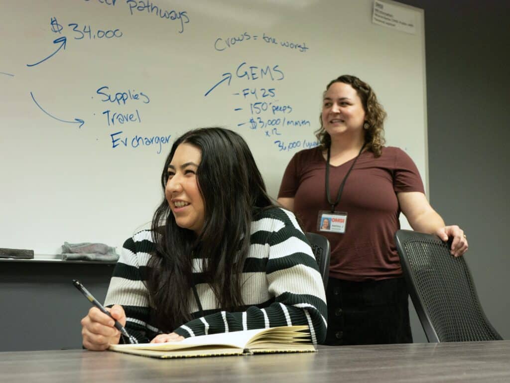 Two OMSI employees smile during a meeting in a conference room. One is sitting and one stands behind her near a whiteboard.