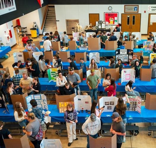 A gymnasium is full of students and tables showcasing their science experiments during the return of the CMHS Science Fair