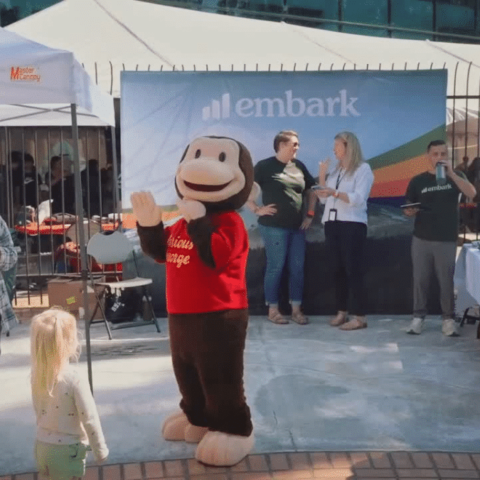 Person in a Curious George costume waving at a little girl at the Oregon Science Festival in front of the Embark table.