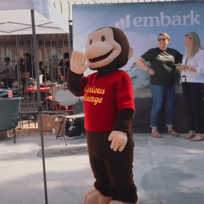 Person in a Curious George costume waving at the Oregon Science Festival in front of the Embark table.