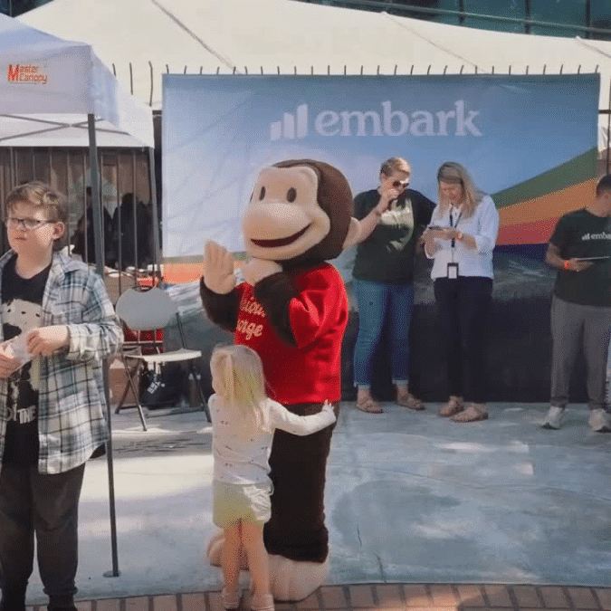 Person in a Curious George costume getting a hug from a little girl at the Oregon Science Festival in front of the Embark table.
