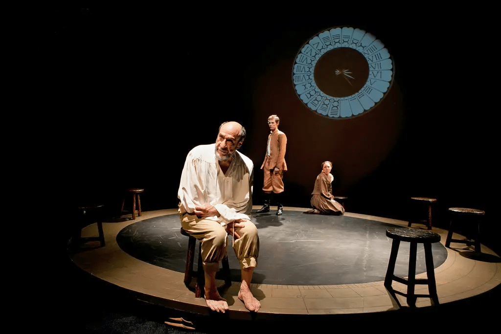 Three actors in worn clothing perform on a dimly lit stage with a circular pattern.