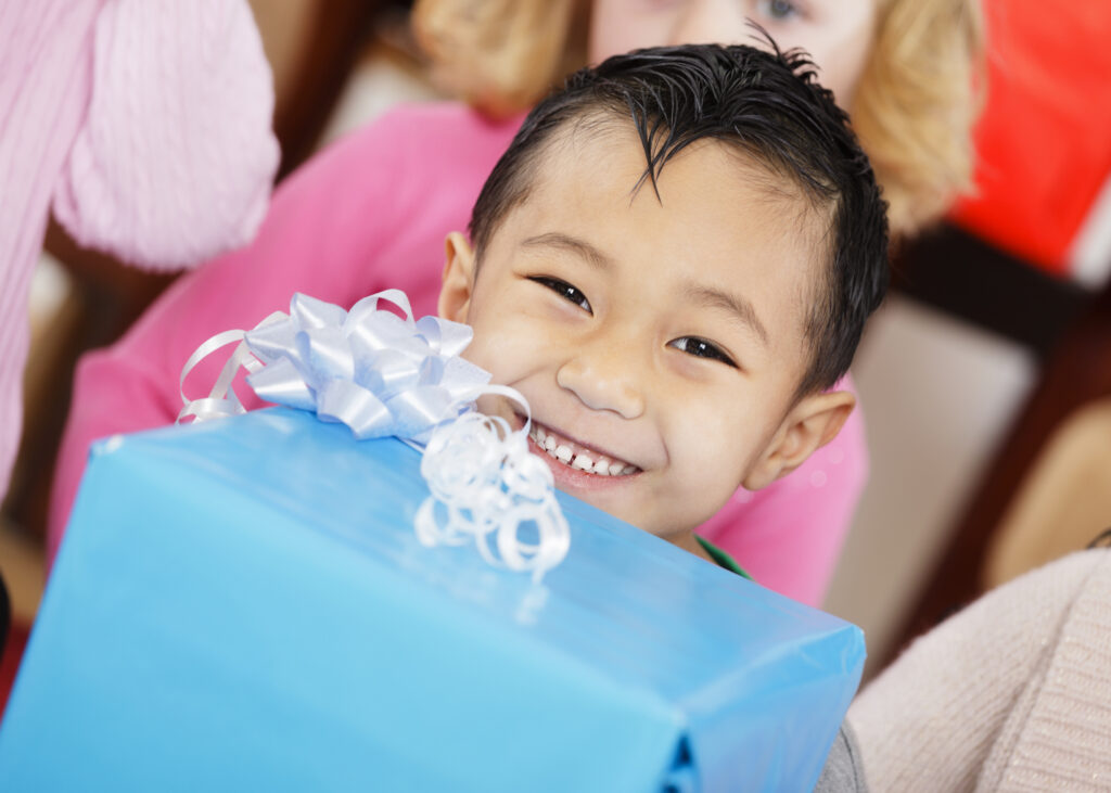 A smiling child holds a blue gift box with a shiny white bow. Other children are visible in the background. The mood is joyful and festive.