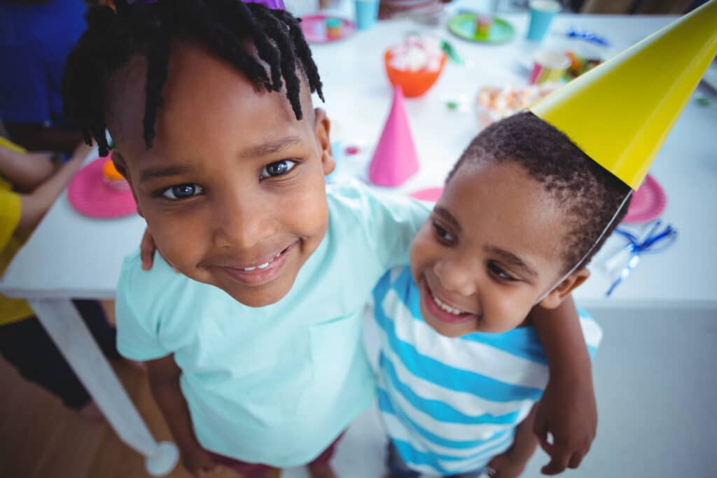 Two smiling children in a birthday party setting, with one wearing a yellow cone hat. They are embracing, surrounded by colorful party decorations. Happy atmosphere.