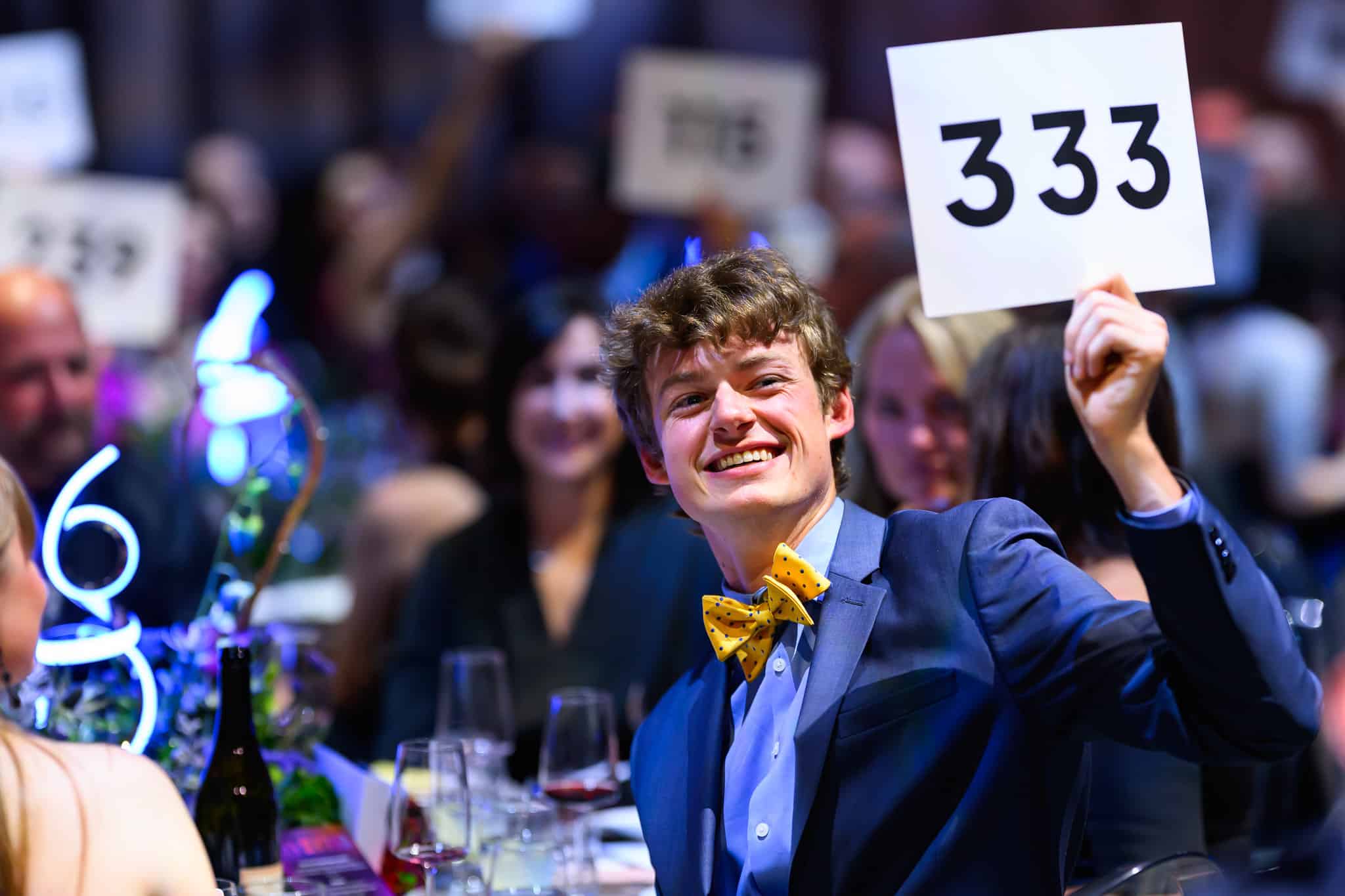 A young man in a blue suit and yellow bow tie smiles, holding up a bidding card with the number 333 at OMSI's annual Gala event.