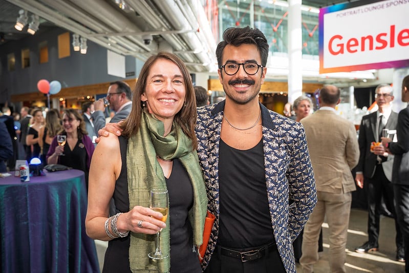 A woman and a man smiling at a lively OMSI Event. The woman wears a green scarf, holding a drink, and the man sports a patterned jacket and glasses.