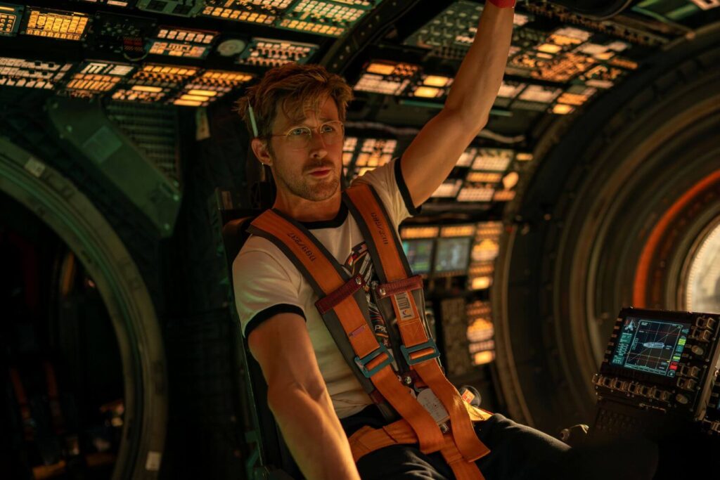 A man with glasses, wearing a white shirt and orange harness, is seated in a futuristic spacecraft cockpit, surrounded by illuminated control panels.