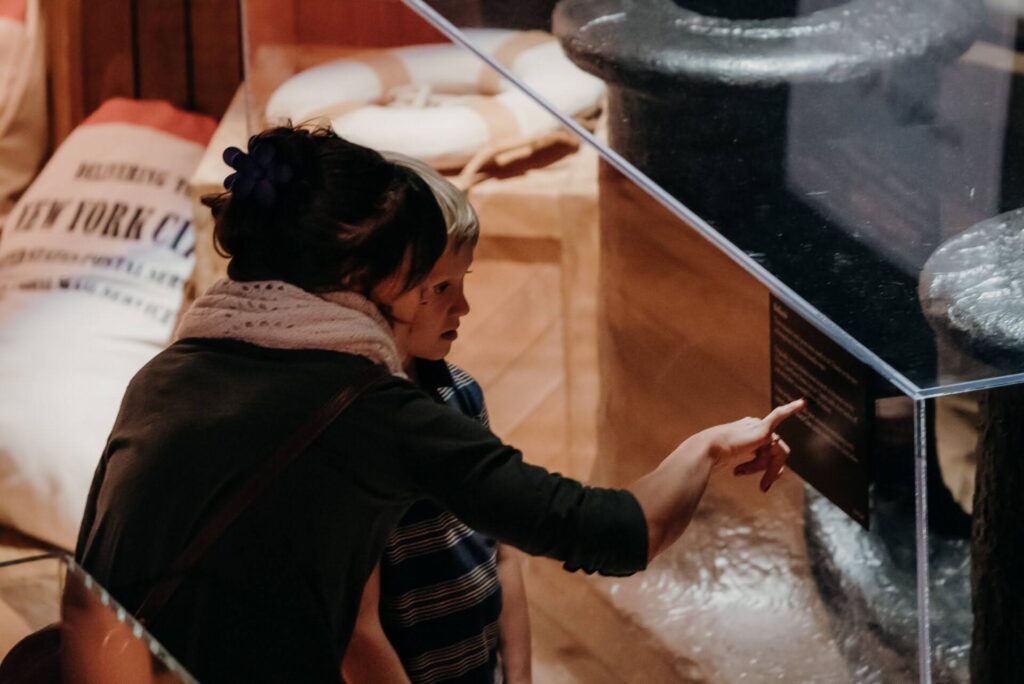 A woman and child in a museum exhibit, engaged and curious. She points at a display case containing historical artifacts. Warm, educational atmosphere.