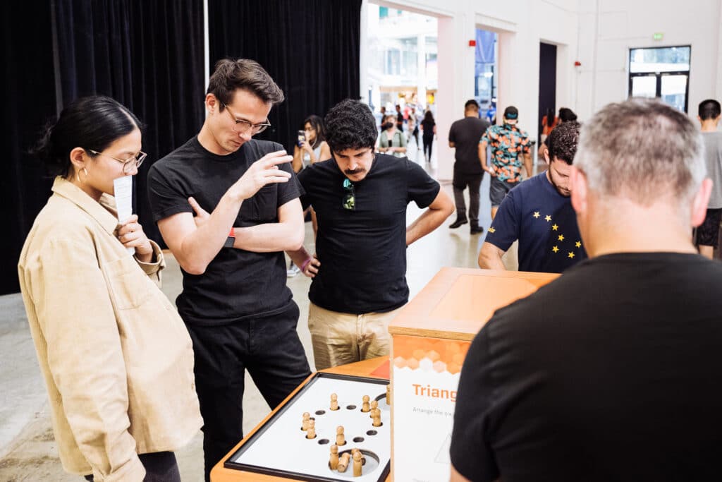 A group of people focuses intently on a tabletop game involving wooden pieces of a brainteaser puzzle at OMSI.