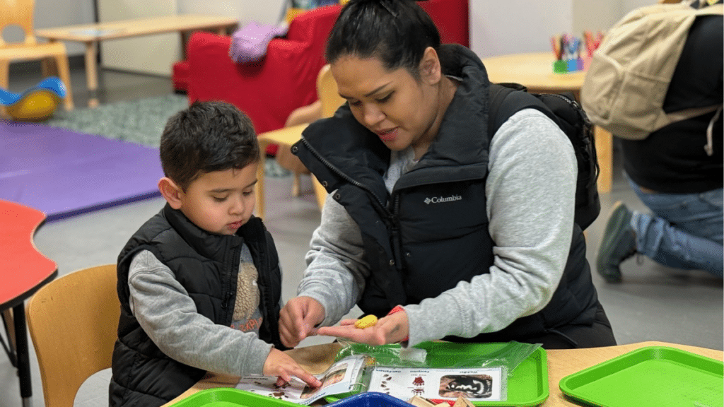 A mother plays with her son at a table with clay and a picturebook.