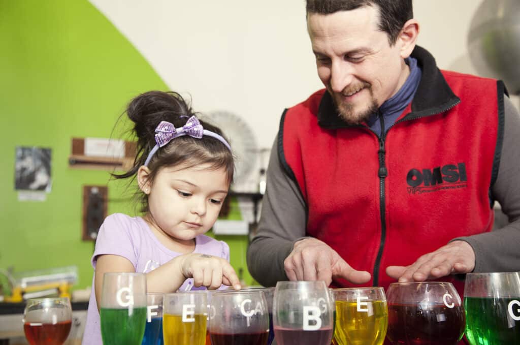 A young girl and an OMSI volunteer learn about frequency and fluid dynamics by running their fingers along the rims of cups filled with different amounts of colored liquid