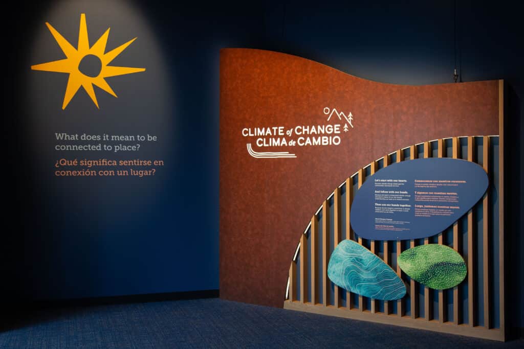 The entrance to the Nancy Stueber Natural Sciences Hall, with the title of the permanent exhibit: Climate of Change / Clima de Cambio. There is a sunburst graphic highlighted to the left and a thank you to sponsors on wooden slats to the right of the image