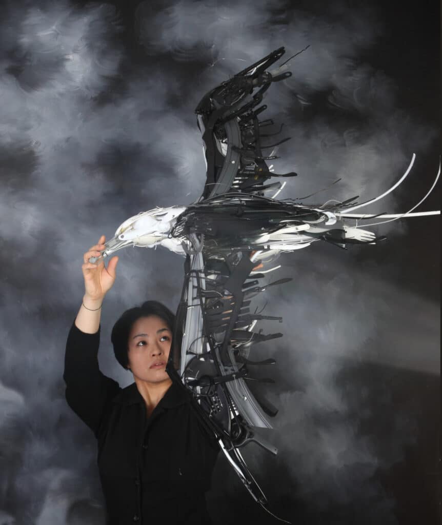 Artist Sayaka Ganz with her piece, Wisdom 2011, Taken by Adam Parsons. It is a large seabird make from black, white, and gray repurposed materials. Ganz is in a black shirt holding up the statue against a black and white cloudy background