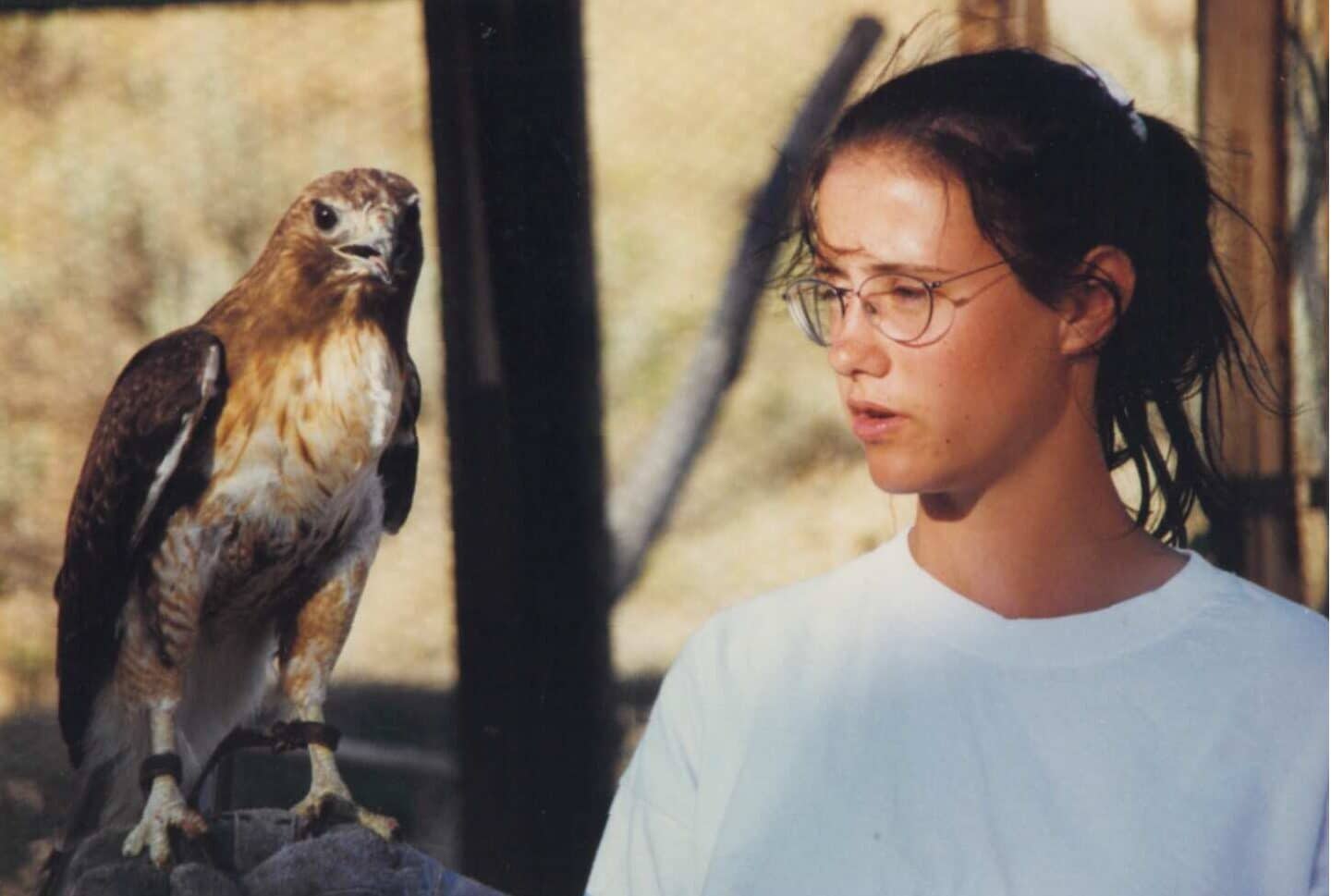 A youth wearing wire-frammed glasses and a white t-shirt holds a falcon on their right arm.