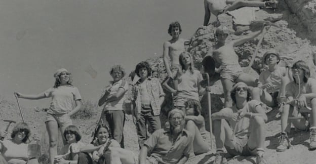 Black and white photo of a group of kids sitting and standing on a rocky hill with shovels and pickaxes