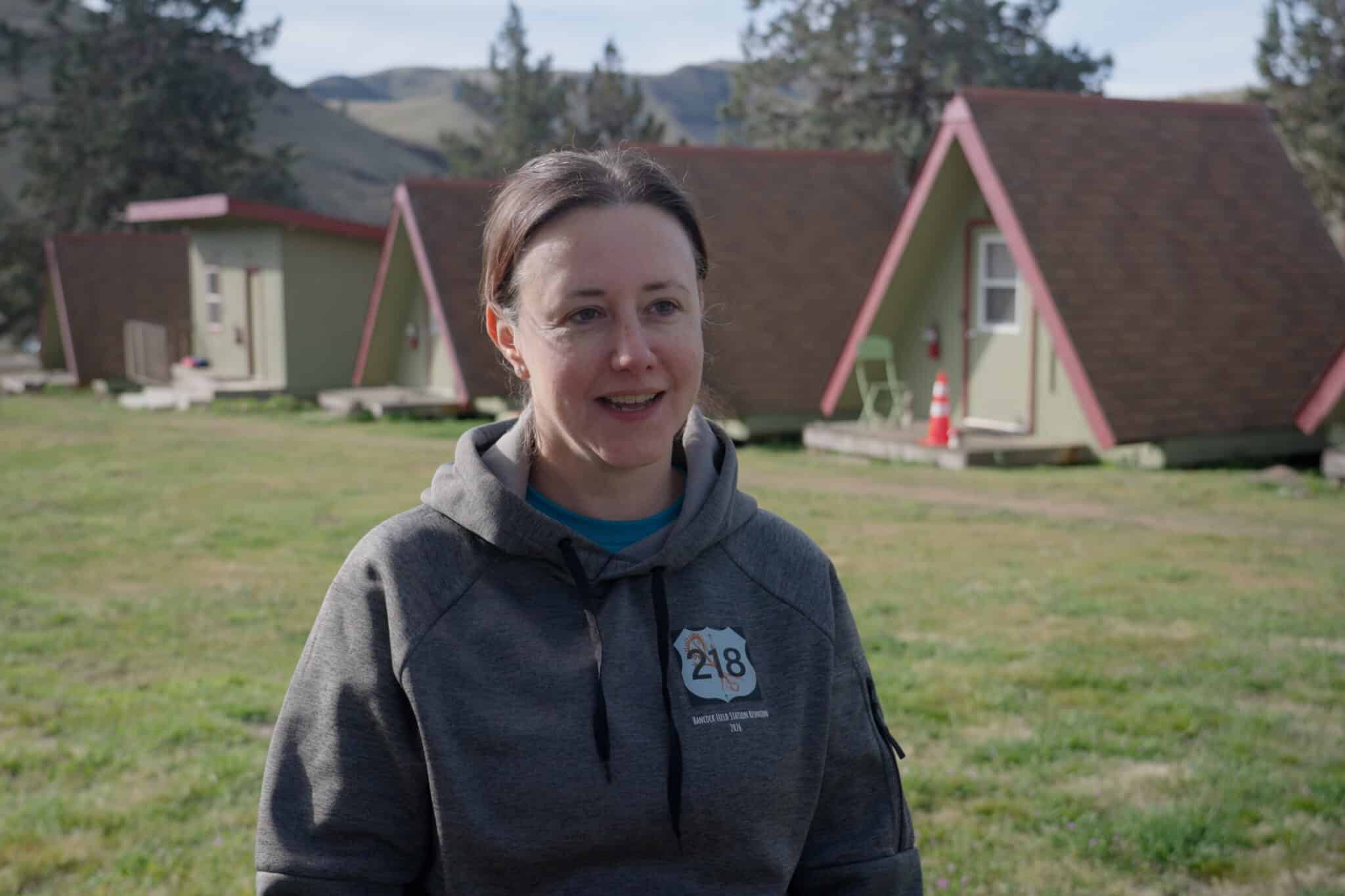 A woman is talking to the camera with Camp Hancock's a-frame cabins in the background