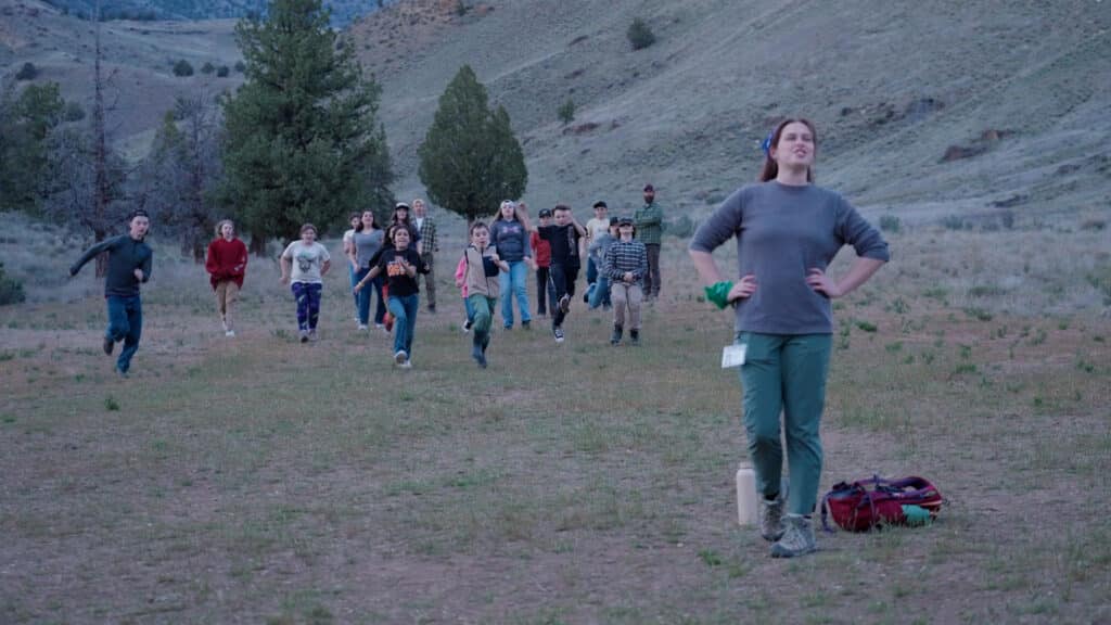 A camp counselor stands facing the camera, with their back turned away from a group of students who are running towards them in a game of Red Light, Green Light in a field at Camp Hancock