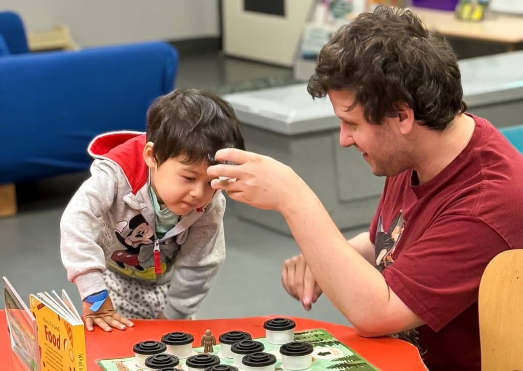 A toddler and an adult are participating in one of the Sasquatch Science Team activities about smell. The adult is holding up a canister and the toddler is smelling it to see if they can identify the contents