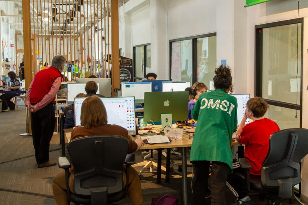 Teens sitting at computers working on design projects with OMSI staff and volunteers in the The Mezz.