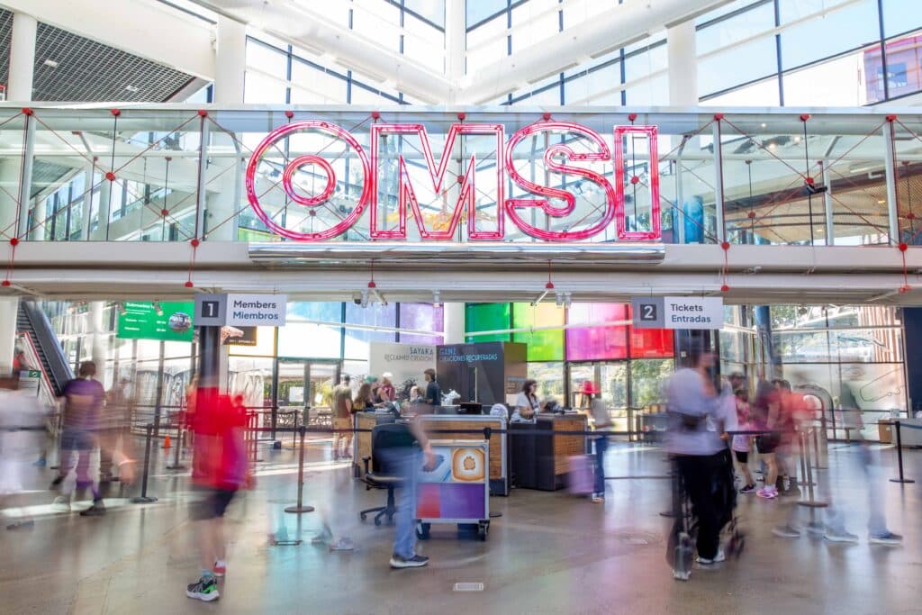 An image of OMSI's lobby and flouescent logo sign. There are blurred visitors in long-exposure motion blur around the base of the image during a busy moment in the museum