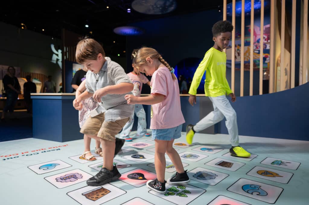 Five kids are playing the floor icon section of Keeping Carbon Stored. The co2 icons are lit up in red and each child is moving to stomp out the co2.