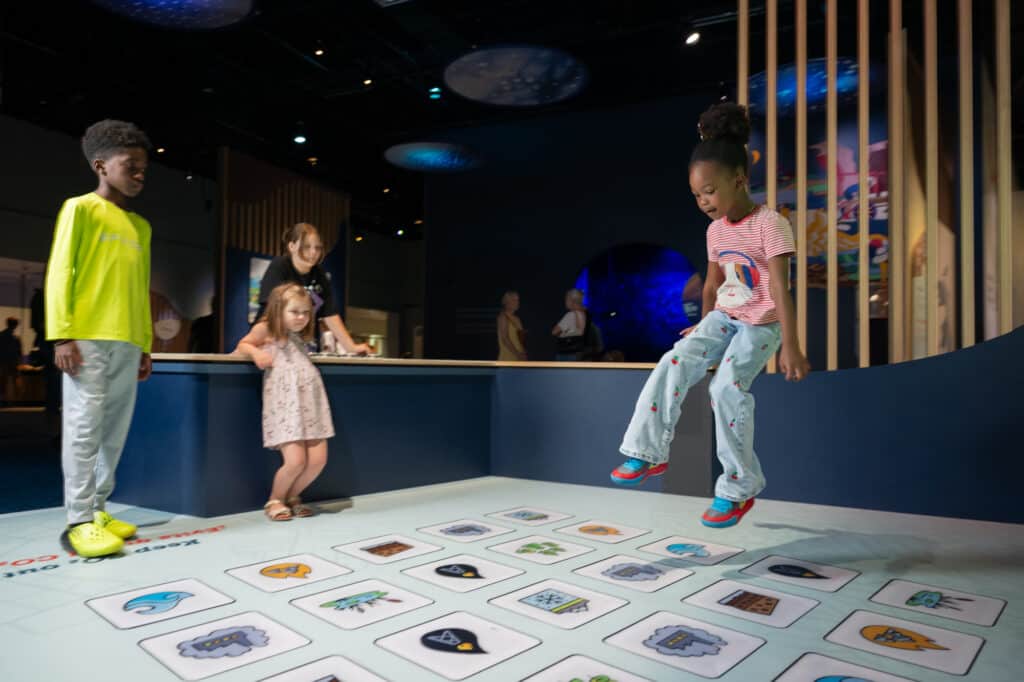 A group of children playing on Keeping Carbon Stored. Of the three kids in the stomping section with floor icons, a young girl is mid-jump and about to land on an icon of carbon.