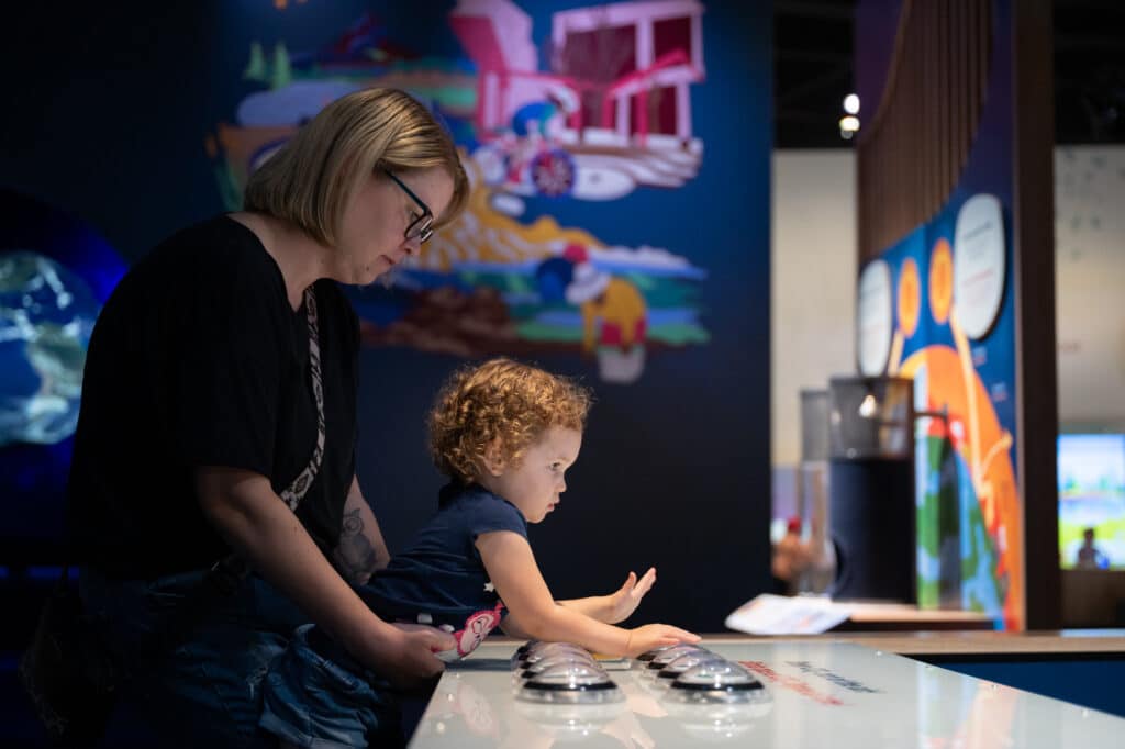 A mother stands behind her child, holding them while they press the buttons on Keeping Carbon Stored. Behind them, the bright colors of a mural stand out against the deep blue wall.