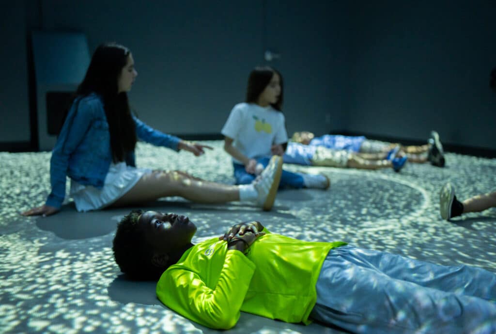 A boy is laying on the ground and looking at the ceiling in the Sit Spot, an immersive installation. Dots of light are being projected across the floor and the boy. Behind him, two girls are sitting on the ground pointing at movement in the light.