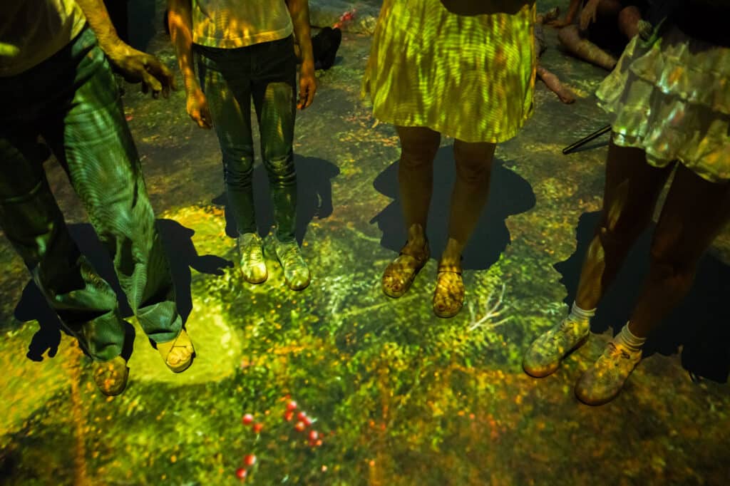 A family stands in the Sit Spot experiences the feeling of walking through a Pacific Northwest forest, including trees, brush, and mushrooms through the projections on the floor.