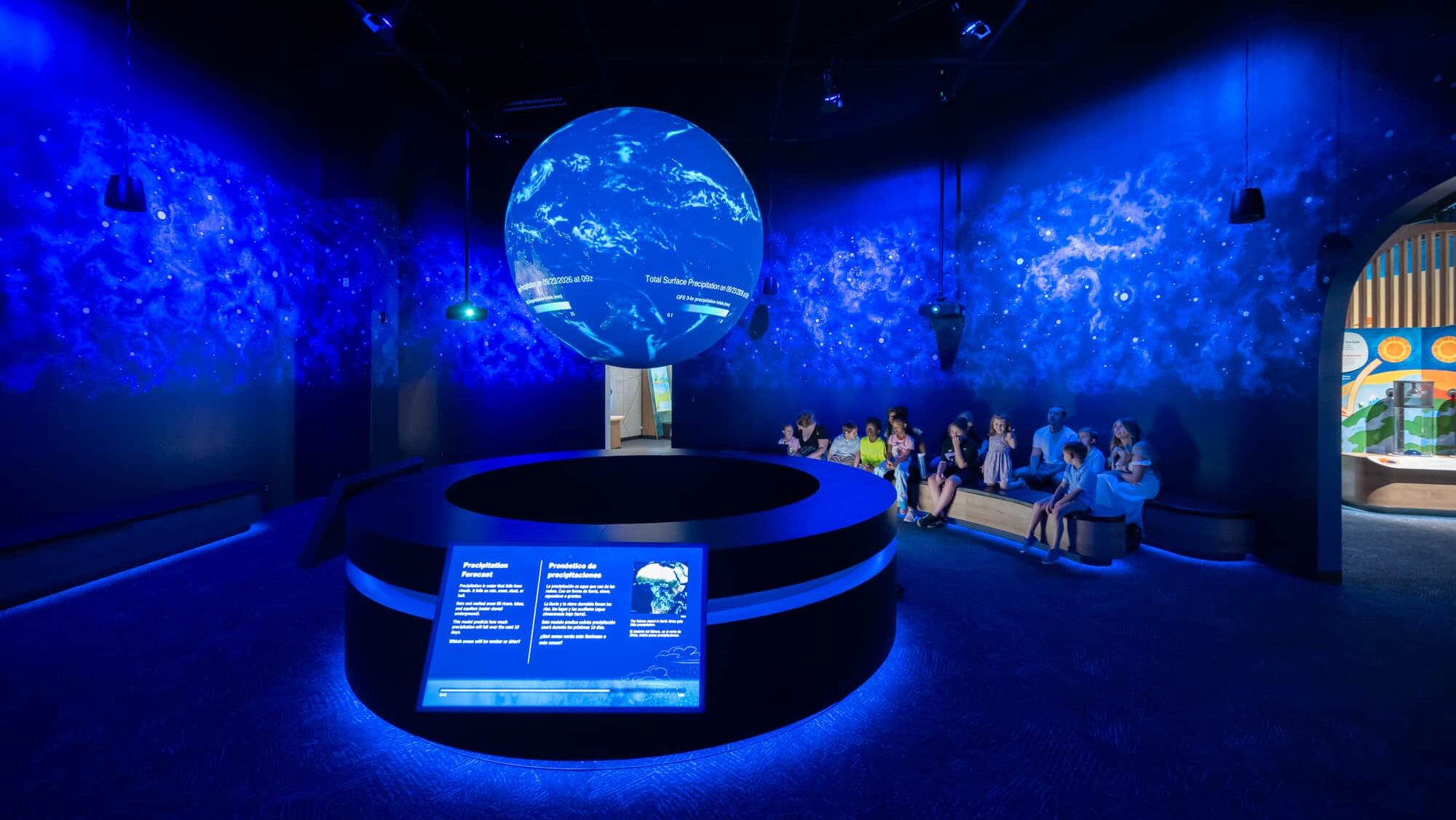 A group of mutliple families sit on benches looking up a the the weather patterns on Science on a Sphere. The sphere looks like it is floating in the middle of the room and is emitting a deep blue light that makes the space themed murals on all the walls stand out.
