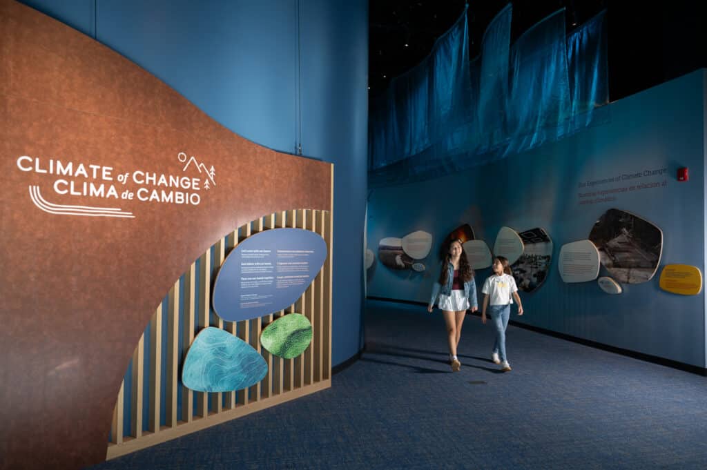 Two not-quite-teenage girls walking out of the Nancy Stueber Natural Sciences Hall with the exhibition logo for Climate of Change | Clima de Cambio in view.