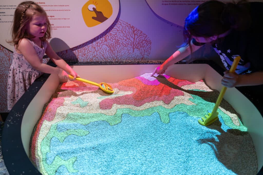 Two children are playing in the Augmented-Reality Sandbox, using yellow tools to mold the sand into various terrain based on a color coded projection onto the sand.