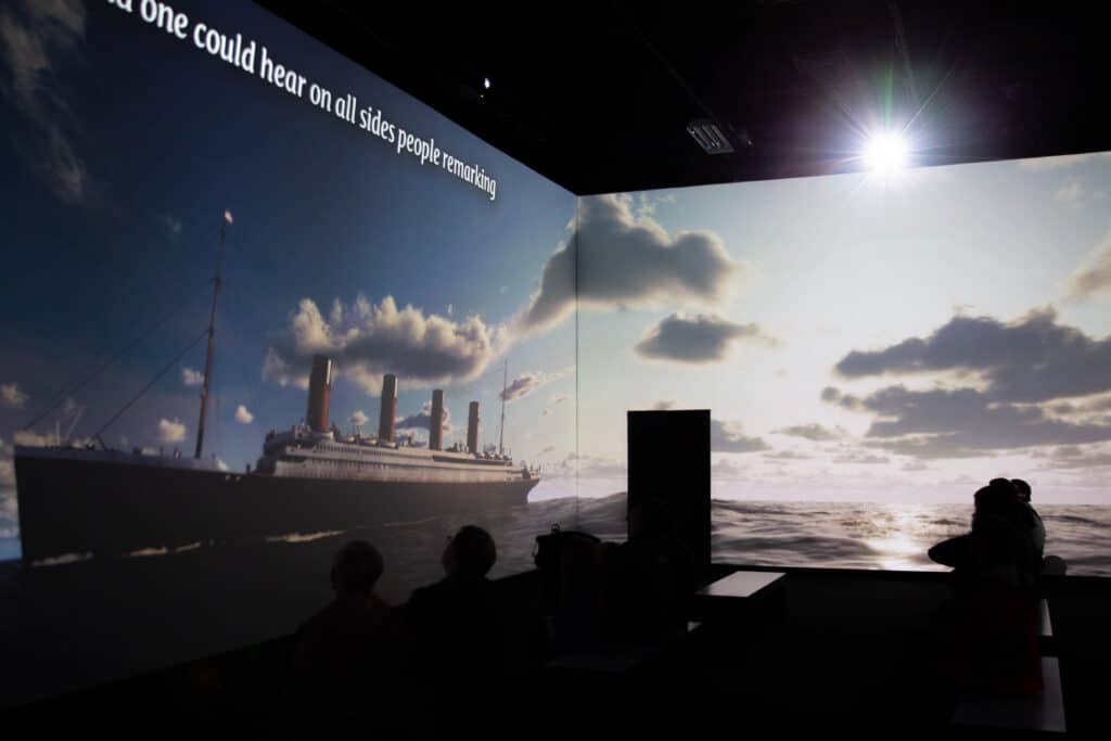 MeyerPro's immersive gallery in TITANIC: The Artifact Exhibition displays the side profile of the Titanic on the ocean in the day. A partially visible quote reads, "one could hear on all sides people remarking."