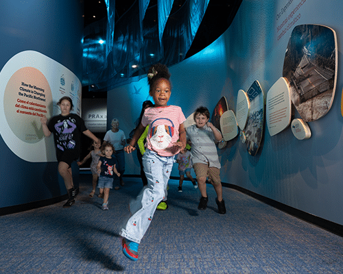An image of several children and adults happily running through the Natural Sciences Hall at OMSI